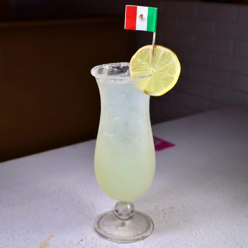 Pale lime cocktail in a salted hurricane glass, garnished with a lime wheel and Mexican flag, sits on a restaurant table.