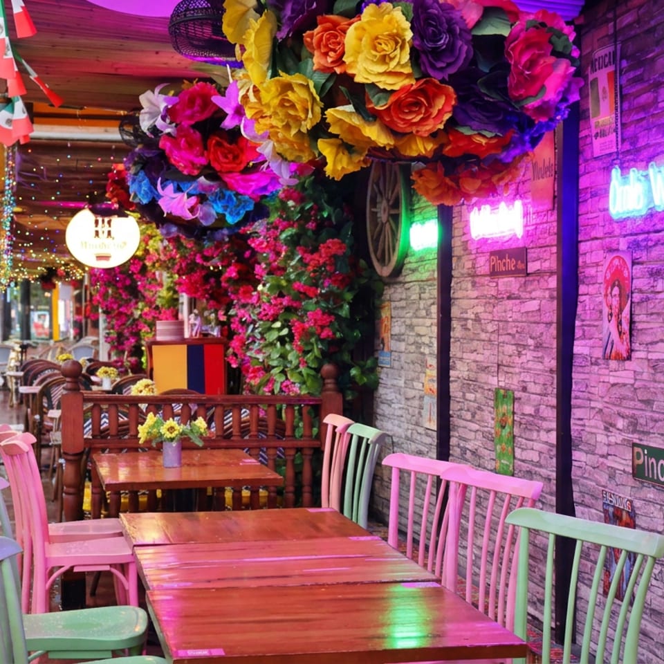 Colorful Mexican restaurant patio with empty wooden tables, pastel chairs, flowers, neon signs, and wet pavement.