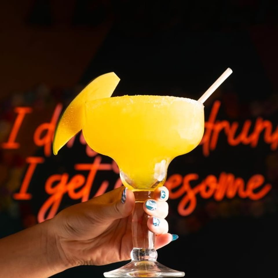 Hand holds a bright yellow frozen cocktail in a margarita glass, garnished with an orange wedge and straw against a neon-lit bar backdrop.