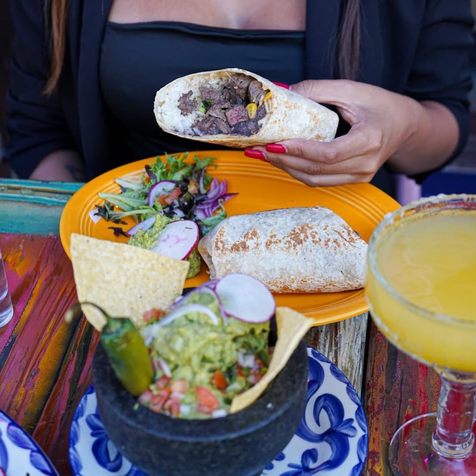 Person holds a steak burrito above a plate with guacamole, salad, tortilla chips, another burrito, and a yellow drink.