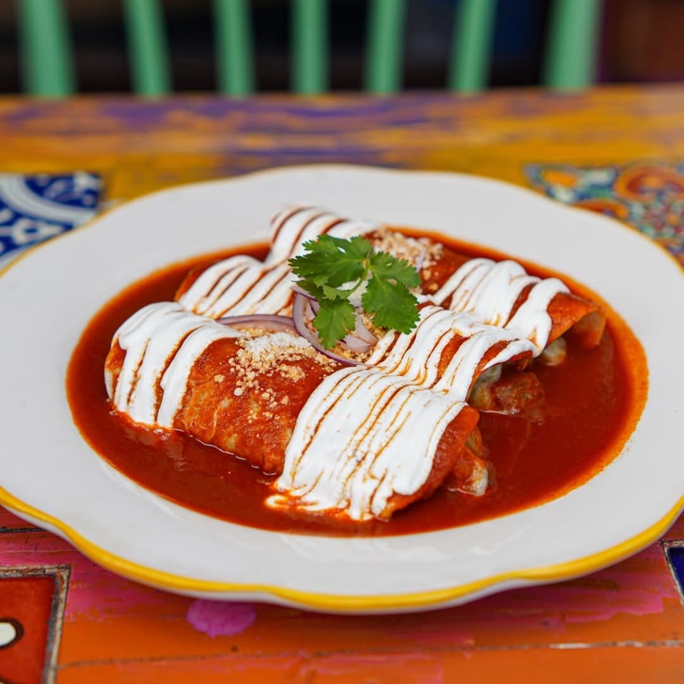 Three enchiladas covered in red sauce and drizzled with crema, topped with cilantro and onions on a colorful plate.