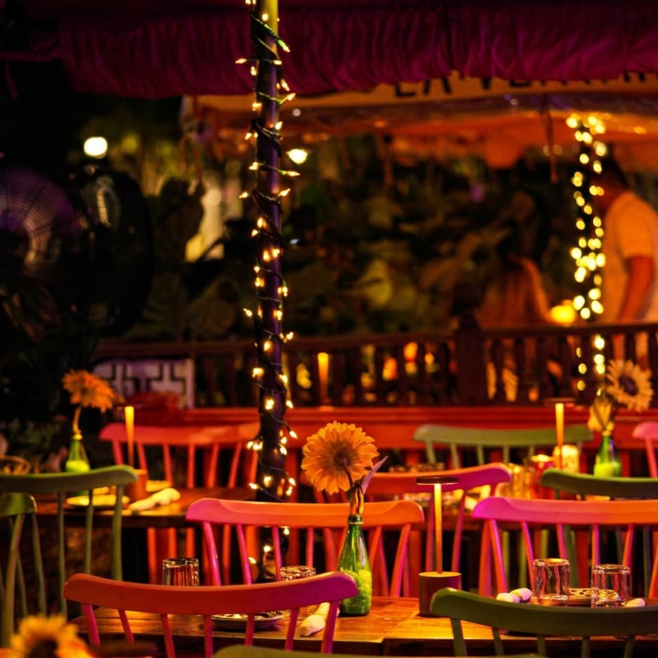 Colorful outdoor restaurant dining area with string lights, wooden tables, painted chairs, and sunflower centerpieces.