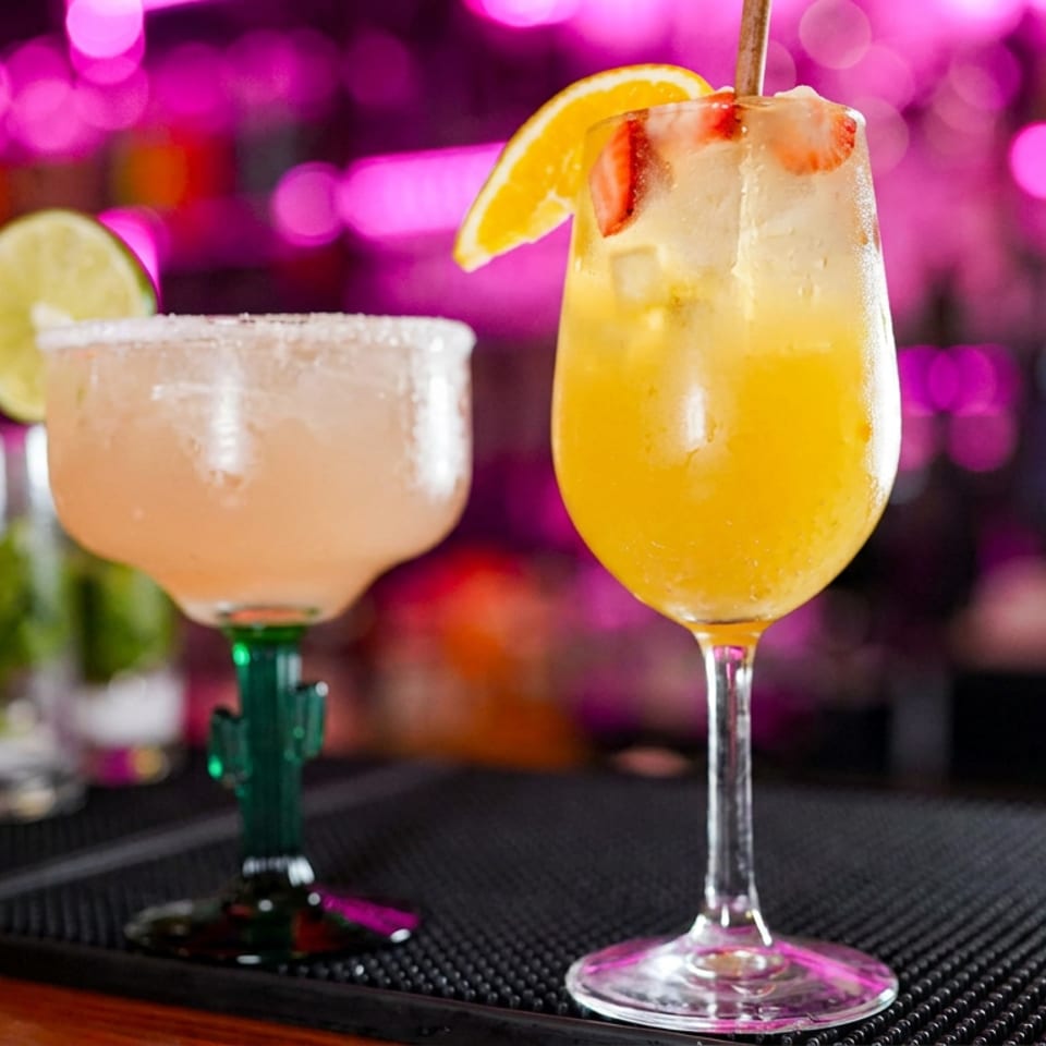 Two colorful cocktails garnished with citrus slices sit on a bar counter against vibrant pink and purple lights.