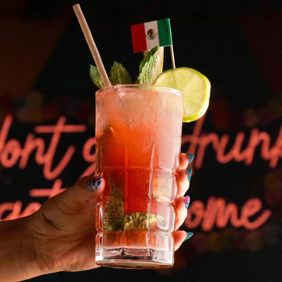 Hand holds a red cocktail garnished with lime, mint, lettuce, and a Mexican flag against a neon-lit restaurant backdrop.