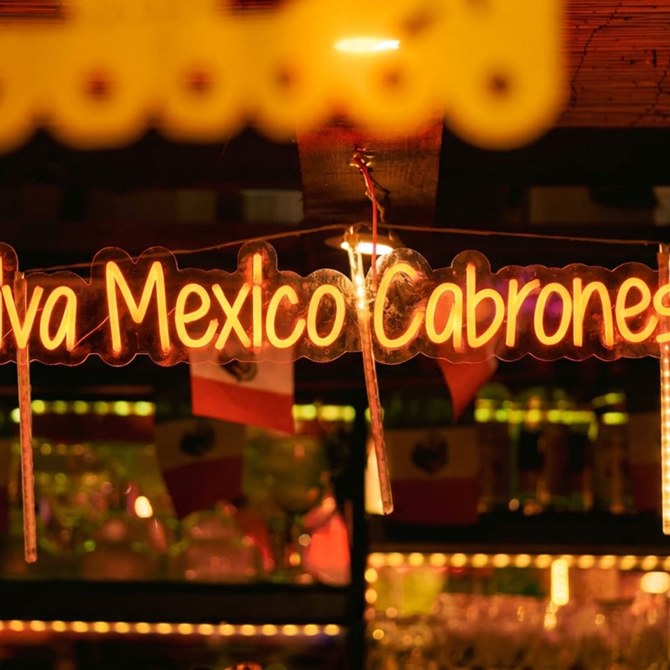Glowing “Viva Mexico Cabrones” sign hangs in a dimly lit Mexican restaurant decorated with flags and warm string lights.