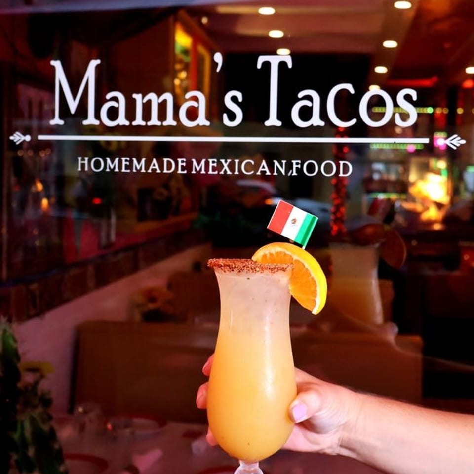 Hand holds a frozen orange cocktail garnished with citrus and a Mexican flag outside Mama’s Tacos, a homemade Mexican food restaurant.