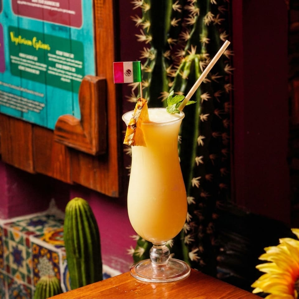 Tropical orange cocktail garnished with pineapple, mint, and a tiny flag, displayed against colorful restaurant decor and sunflowers.