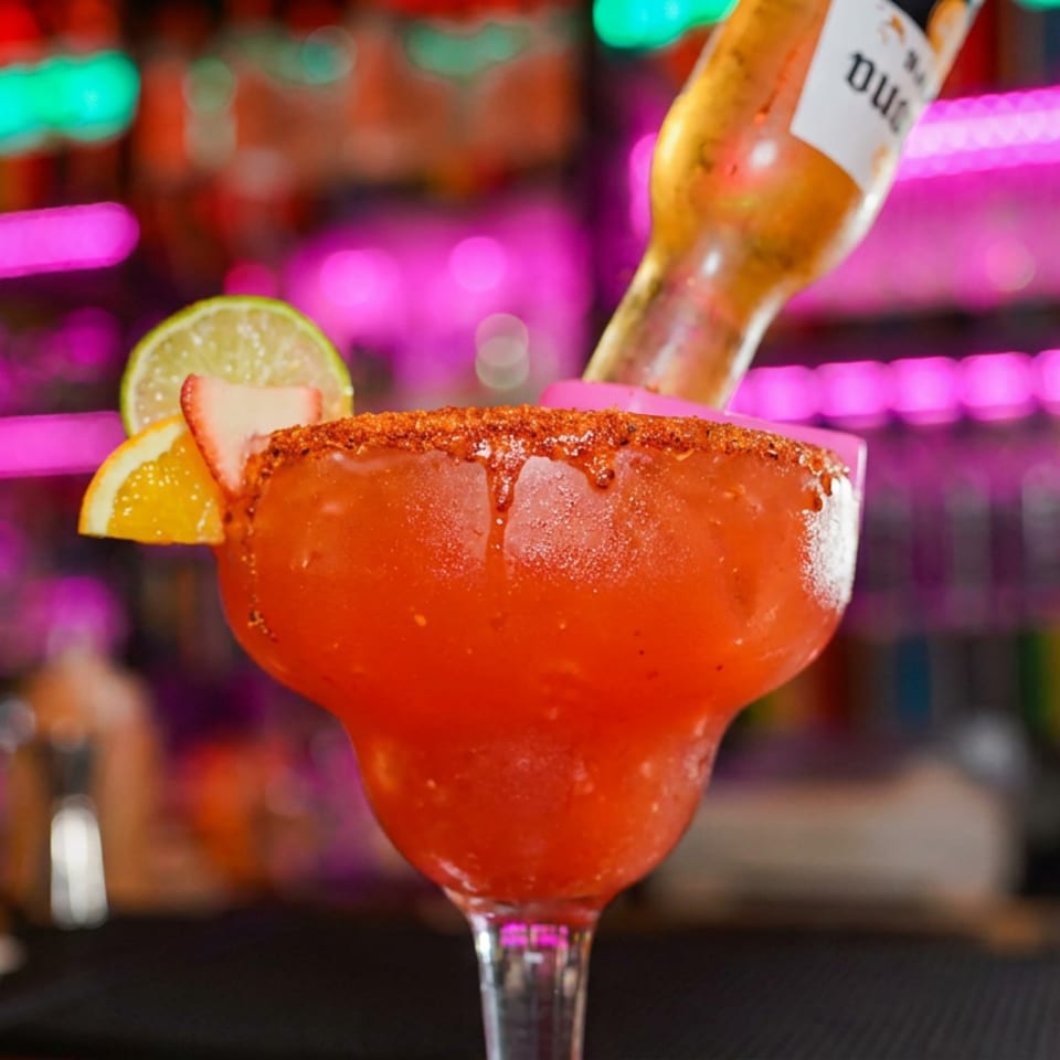 Red frozen cocktail with chili-salt rim, citrus garnish, and bottle behind it on a colorful neon-lit bar.