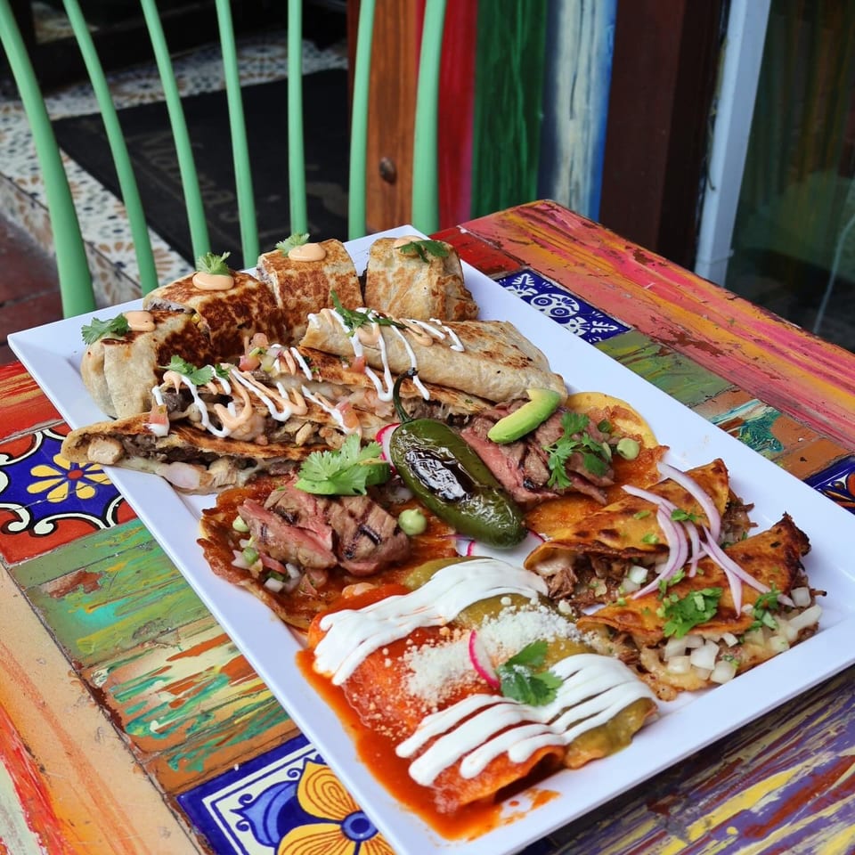 Assorted Mexican dishes with grilled meat, quesadillas, tortillas, creamy sauces, cilantro, and roasted jalapeño on a colorful tiled table.