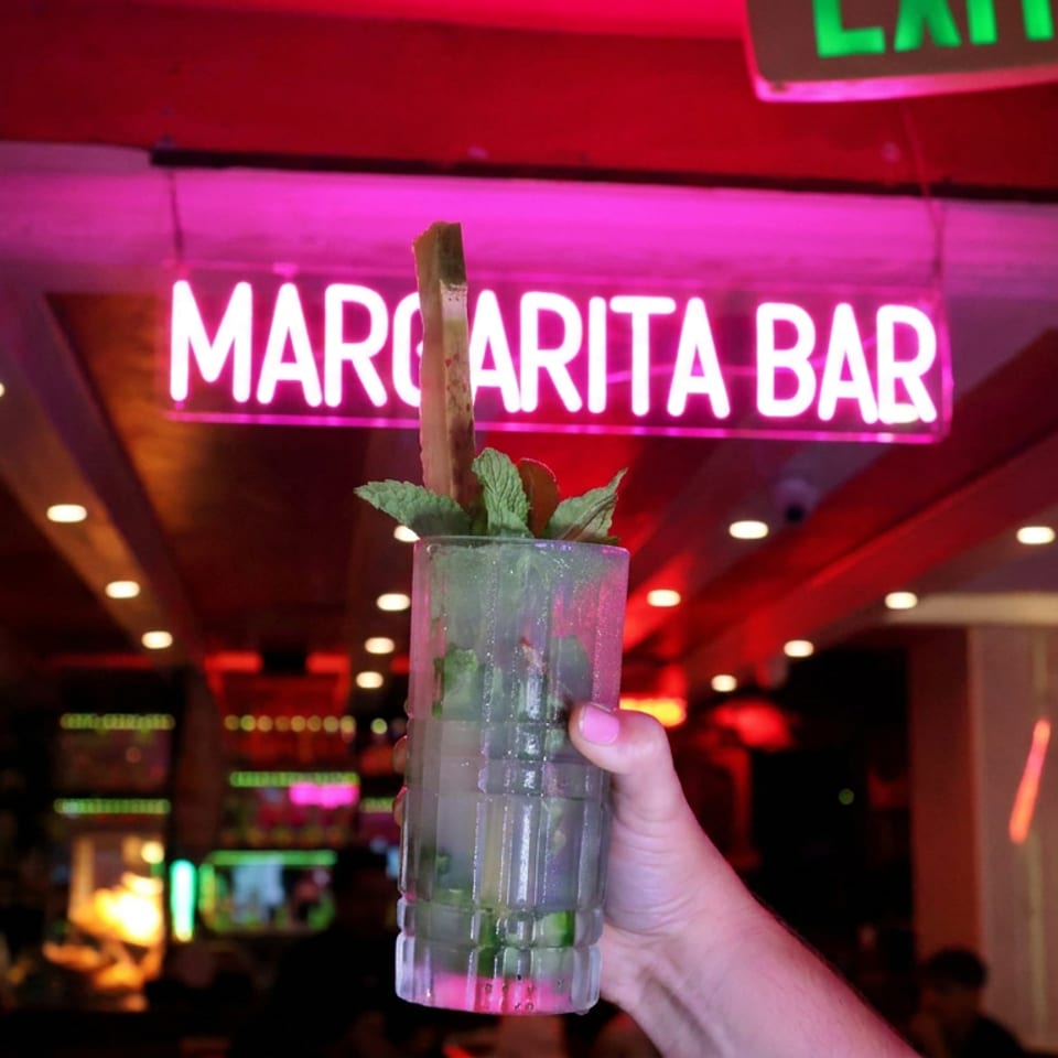Hand holds a mint-garnished cocktail before a glowing Margarita Bar sign in a vibrant, red-lit restaurant.