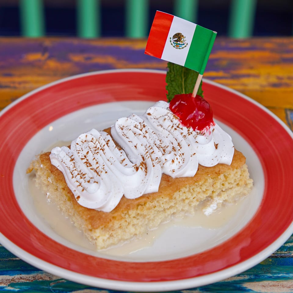Mexican tres leches cake topped with whipped cream, cinnamon, cherry, and a small Mexican flag on a red-rimmed plate.