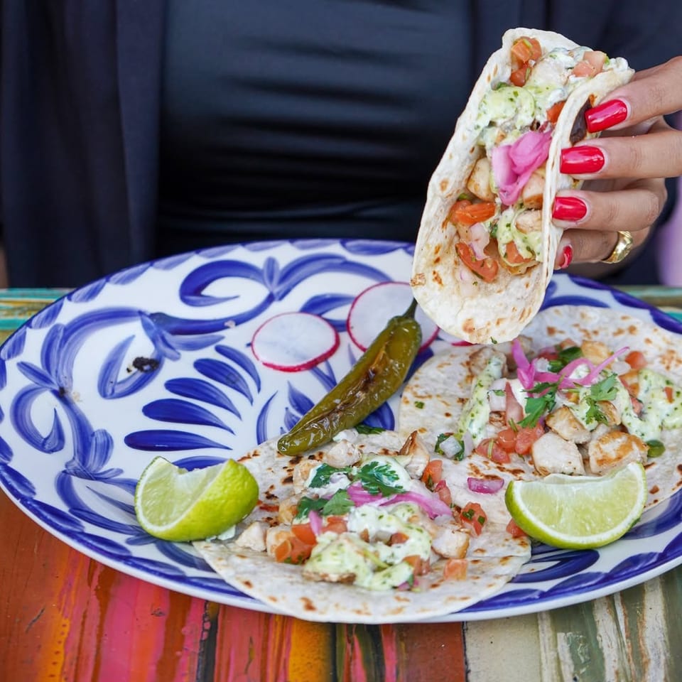 Hand holds a chicken taco over a colorful plate of tacos garnished with lime, radish, pickled pepper, and fresh toppings.