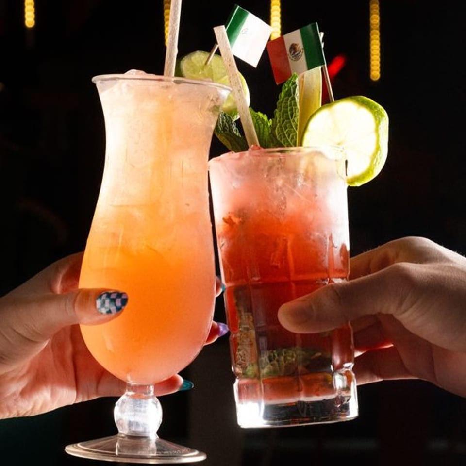 Two colorful cocktails garnished with lime, mint, and Mexican flags are raised in front of a glowing “Viva Mexico Cantina” sign.