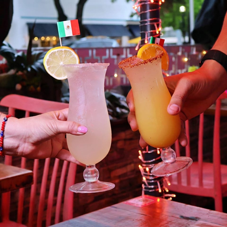 Two colorful cocktails with citrus garnishes are held in a festive outdoor restaurant patio decorated with string lights.