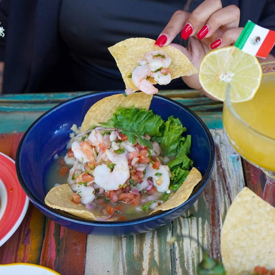 Bowl of shrimp ceviche with tortilla chips and lettuce, while a hand holds a chip beside a lime-garnished citrus drink.